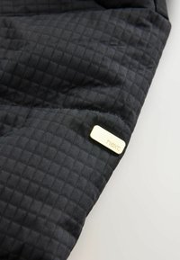 Tessut negru matlasat cu un model texturat, având eticheta cu logo-ul „next” de culoare aurie metalizată. Tesătura are o finisare netedă, mată.