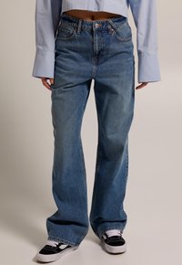 Light blue, high-waisted denim jeans with a wide-leg cut, featuring classic pockets and seam detailing. Paired with black and white sneakers.