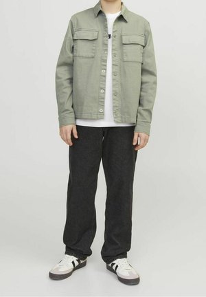 Henkilö, joka käyttää vaaleanvihreää napillista takkia, valkoista t-paitaa, löysiä mustia housuja ja valkoisia tennareita, joissa on mustat raidat, seisoo vaalealla taustalla.