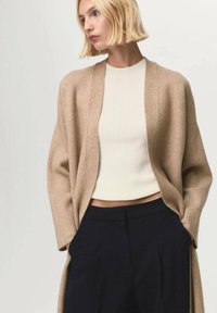 Donna bionda che indossa un cardigan aperto beige, un top corto a coste bianco e pantaloni neri a vita alta, mentre guarda di lato.