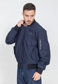 Marineblauer Bomberjacke mit Reißverschluss vorne, gerippten Bündchen und zwei Seitentaschen. Verfügt über eine zusätzliche Reißverschlusstasche am linken Ärmel für zusätzlichen Stauraum.