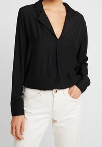 Blouse noire à manches longues avec un col en V, présentant un décolleté plongeant et une coupe décontractée, associée à un jean blanc taille haute.