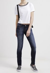 Camiseta corta blanca con ribete negro, combinada con jeans skinny azul oscuro y zapatos gris sin cordones. Modelo de pie contra un fondo liso.