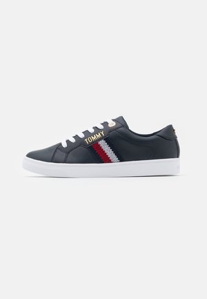 Baskets basses noires avec semelle blanche, lacets blancs, détail en tissu rayé rouge, blanc et marine, et logo doré « TOMMY » sur le côté.