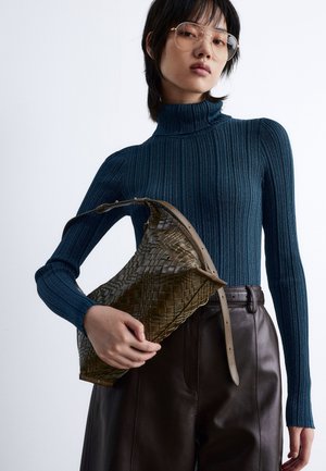 Green woven leather bag with a curved design, paired with a ribbed dark blue turtleneck and high-waisted brown leather pants.