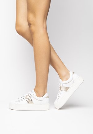 Baskets blanches avec des accents métalliques dorés, une semelle épaisse, un design à lacets et une texture douce, présentées de côté avec des jambes partiellement visibles.