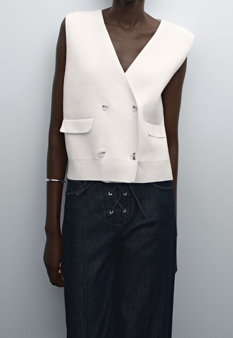 Biała, bezrękawnikowa kamizelka z podwójnym rzędami guzików i kieszeniami, noszona na ciemnych, wysokich spodniach dżinsowych z wiązaniem z przodu, ozdobiona srebrną bransoletką.