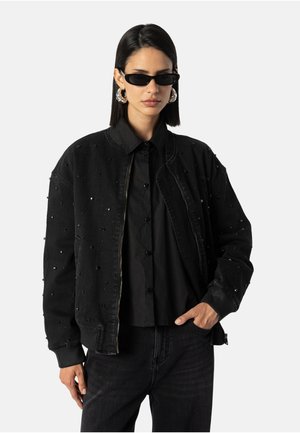 Schwarze Jeansbomberjacke mit verzierten Perlendetails, gerippten Bündchen und Kragen; getragen über einem schwarzen Hemd mit Knöpfen, kombiniert mit Sonnenbrille.