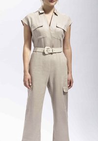 Žena u bež kratkom kombinezonu s džepovima na prsima, širokim remenom s pravokutnom kopčom i hlačnim džepovima u cargo stilu, stoji ispred jednobojne pozadine.