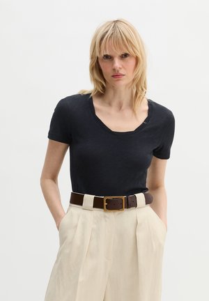 Blonde vrouw die een zwart T-shirt met korte mouwen draagt, in een hooggetailleerde beige geplooide broek met een donkerbruine riem, handen in de zakken.
