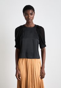 Top en satin noir avec des manches bouffantes en tulle, associé à une jupe plissée beige. Encolure simple et coupe ajustée mettent en valeur la silhouette.