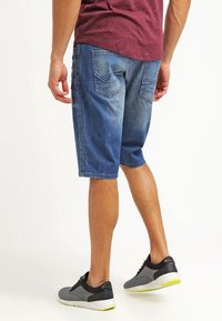 Džínové šortky středně modré barvy s ošoupaným vzhledem, mají dvě zadní kapsy a rovný střih, v kombinaci s černými a šedými sportovními botami.