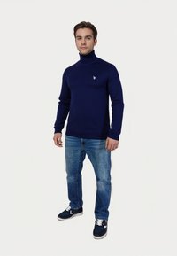 Hombre vestido con un suéter de cuello alto azul marino, jeans azules y zapatillas azul marino, de pie frente a un fondo blanco liso.