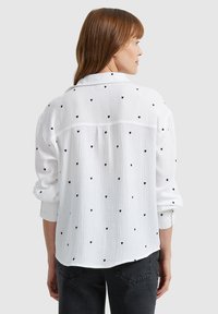 White button-up shirt made of textured fabric, featuring black heart patterns and a relaxed fit with rolled sleeves.