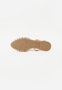 Roze gouden sandaal met een gevlochten bandontwerp, getextureerde rubberen zool en gegraveerd merklogo. Plat met een ronde neus en minimale details.