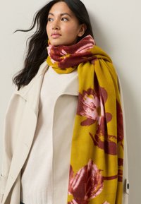 Pehme kollane sall roosa ja bordoopunase lillemustriga, kaela ümber heidetuna, sobitatuna kreemja kampsuni ja helebeeži mantliga.