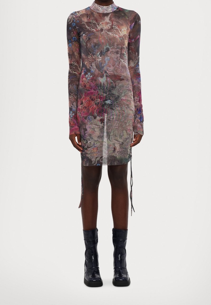 Robe ajustée à manches longues avec motif abstrait multicolore, associée à des bottines noires brillantes portée par une personne debout devant un fond uni.
