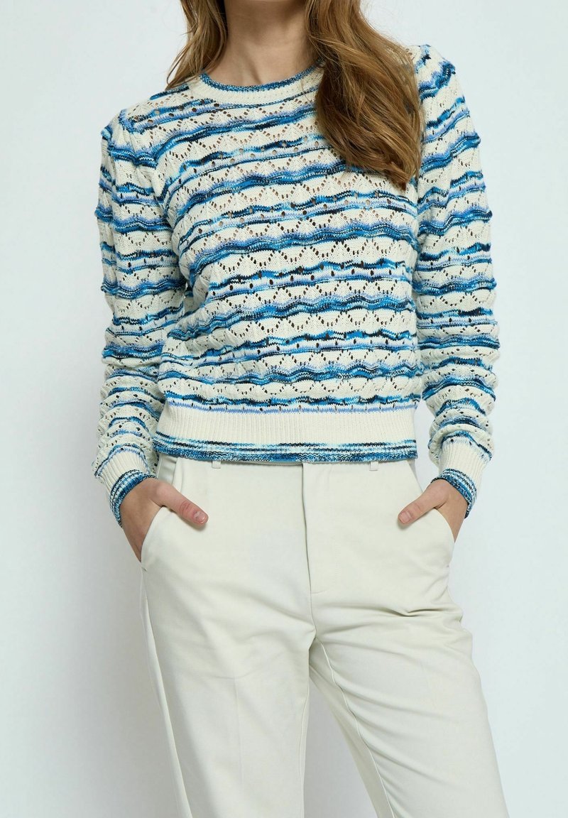 Sweter robiony na drutach z teksturowanym wzorem fali w kolorze niebieskim i kremowym, z okrągłym dekoltem i ściągaczami na rękawach, w zestawieniu z jasnobeżowymi spodniami.