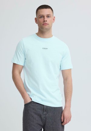 Mand med kort hår iført en lyseblå T-shirt med et lille sort logo på brystet og mørkegrå jeans, stående med den ene hånd i lommen.