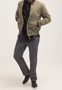 Giacca bomber verde oliva, con zip frontale, abbinata a una camicia nera con bottoni e pantaloni grigi. Sneakers nere con suole bianche.