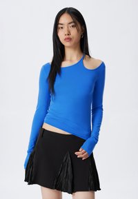 Junge Frau mit langen schwarzen Haaren, die ein leuchtend blaues, einseitiges Langarmoberteil und einen schwarzen Rock mit gefältelten Paneelen trägt.
