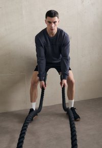 Schwarze Battle Ropes werden von einer Person in einem dunklen Sweatshirt und schwarzen Shorts gehalten, die auf einem grauen Boden gegen eine strukturierte beige Wand steht.