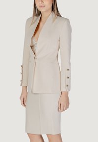 Blazer beige con rever appuntito, chiusura a bottoni e quattro bottoni dorati su ciascun maniche. Abbinato a una gonna beige aderente.