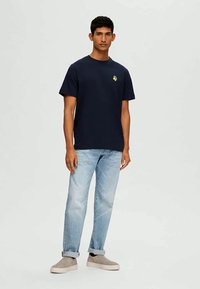T-shirt de algodão azul-marinho com mangas curtas e um gráfico amarelo no peito, combinada com jeans azul-claro e sapatos de slip-on bege.