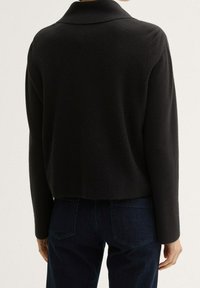 Černý texturovaný krátký cardigan s límečkem, dlouhými rukávy. V kombinaci s tmavě modrými džíny, viděno z zadní strany.