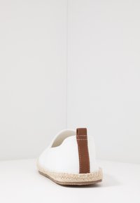 Espadrille en toile blanche avec une semelle en jute tressé et un onglet de talon en cuir marron contrastant. Conception à enfiler, couture minimale visible.