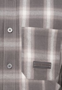 Chemise à carreaux gris et blanc à l'avant avec des boutons et une poche avec une étiquette de marque CAIX cousue.