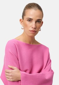 Pull en maille côtelée rose avec des manches larges et évasées. Le mannequin pose les bras croisés, mettant en valeur la texture et la couleur du vêtement.