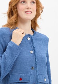 Blau strukturierte Jacke mit einem ausgefransten Ausschnitt, silbernen Knöpfen und einem roten Akzentknopf, ausgestattet mit langen Ärmeln und aufgesetzten Taschen.