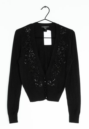 Czarny kardigan z długim rękawem na przód zapinany na guziki, ozdobiony kwiatowymi koralikami i cekinami wzdłuż dekoltu, zawieszony na czarnym wieszaku z białą metką.