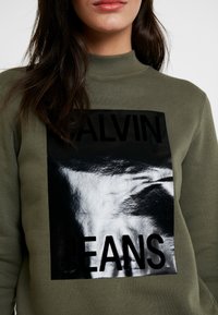 Sudadera verde oliva con un cuello alto, que presenta un gran gráfico en negro y plateado brillante con la inscripción "CALVIN JEANS."