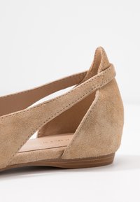 Beżowe zamszowe baleriny z niskim obcasem w kształcie koturny, z wyciętymi bokami i teksturowaną powierzchnią. Wnętrze zdobi złoty napis marki.