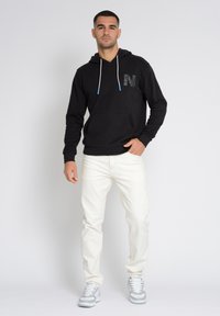 Musta huppari, jossa on kuvioitu tasku ja sinisin kärjin varustettu nyöri. Yhdistettynä valkoisiin farkkuihin ja harmaisiin urheilukenkäihin. Minimalistinen muotoilu.