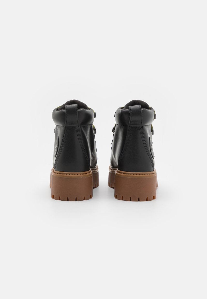 Stivaletti neri in pelle con punta arrotondata, suola in gomma marrone spessa e accenti in tessuto verde. Presentano doppi cinturini laterali e superficie testurizzata.