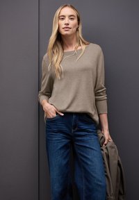 Maglione a maglia beige con maniche lunghe, abbinato a jeans a gamba larga blu scuro. Il modello tiene una giacca grigia, in piedi su uno sfondo grigio.