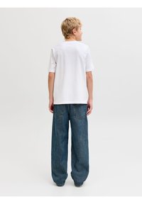 Ξανθό παιδί στέκεται με την πλάτη, φορώντας ένα απλό λευκό κοντομάνικο πουκάμισο και φαρδιά μπλε τζιν, με φόντο ένα λευκό τοίχο.