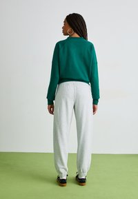 Sudadera verde corta, pantalones de chándal grises, puños elásticos, zapatos planos. Cabello trenzado, grandes pendientes de aro, fondo liso. Texturas suaves visibles.