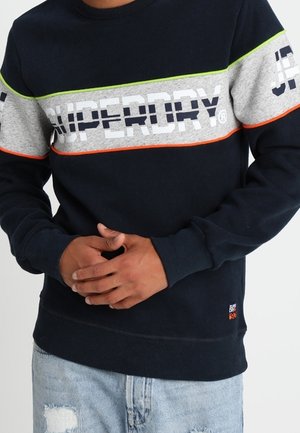 Mann trägt marineblaue und graue Superdry-Sweatshirt mit orangenen und grünen Streifen, Hände vor dem Körper verschränkt, kombiniert mit hellen, zerrissenen Jeans.
