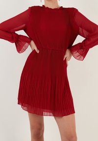 Abito rosso a pieghe con maniche lunghe e trasparenti e scollatura arricciata. Il tessuto è leggero e fluisce dolcemente, creando una texture simile a onde.