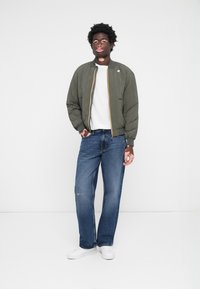 Olíva zöld, steppelt bomber dzseki sárga cipzárral, fehér póló és laza farmernadrág. A fehér sportcipők teszik teljessé az outfitet.