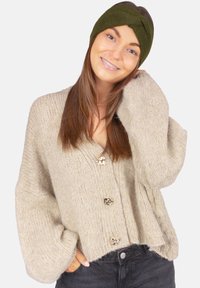 Cardigan beige tricoté avec manches surdimensionnées et boutons tête de lion dorés, associé à un serre-tête vert foncé. La texture semble douce et épaisse.