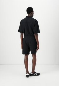 Sort jumpsuit med korte ærmer, elastisk talje og baglomme, lavet af tekstureret stof, parret med sorte træningssko med hvide detaljer.