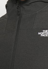 Giacca nera con zip in tessuto testurizzato, dotata di un colletto alto. Presenta un logo bianco prominente sul petto e dettagli in zip.
