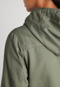 Närbild av en persons axel och nacke som bär en grön huva med tydliga sömnadsdetaljer.