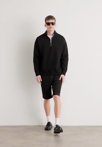 Sweat à zip noir, shorts noirs, chaussures noires, chaussettes blanches ; le modèle porte des lunettes de soleil ; design minimaliste sur fond uni.