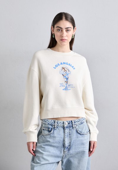 Sweatshirt court crème avec un graphique et une illustration bleus "Los Angeles". Présente des épaules tombantes et une texture de tissu douce.